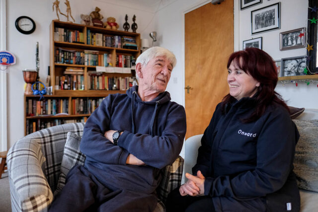 An Onward employee and a customer sit inside the customer's living room, laughing in conversation. They sit on comfortable armchairs, with a large bookshelf behind them.